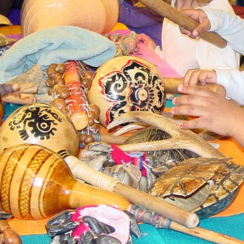 Three young girls touch and examine colorful painted musical instruments made from plant and animal materials at a presentation table.