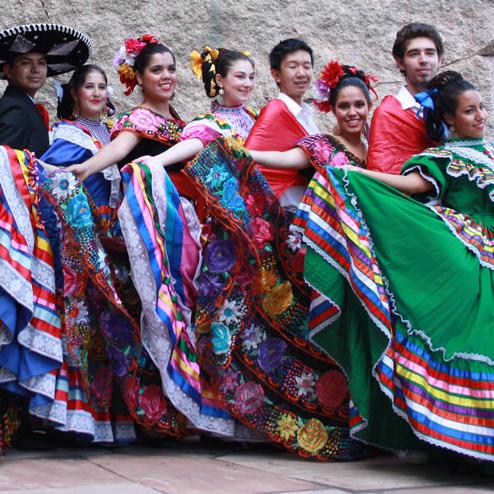 Eleven smiling dancers in colorful traditional Mexican dress pose along the stone base of the Peabody’s outdoor Torosaurus sculpture. 