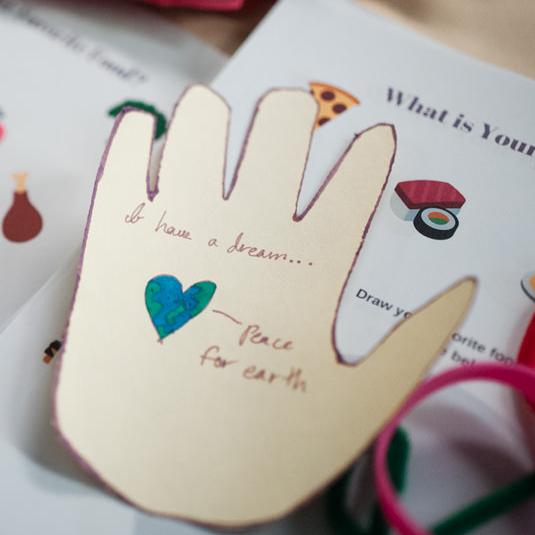 Someone’s hands decorate a cut-out of a hand at a craft table piled high with waiting paper hands.