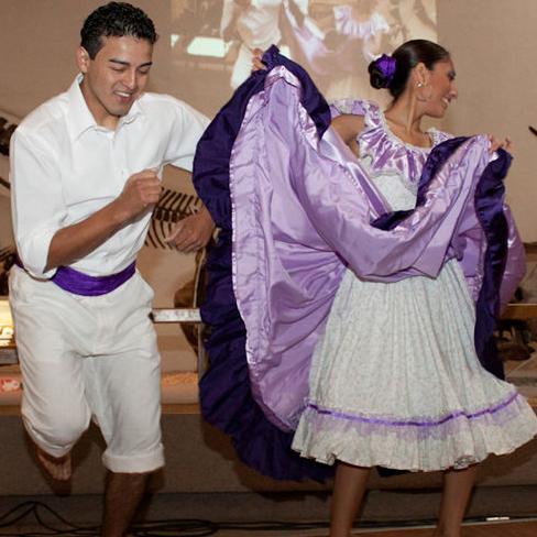 A woman in a purple traditional dress and her male partner dance flamboyantly barefoot in front of the Peabody dinosaur skull display.