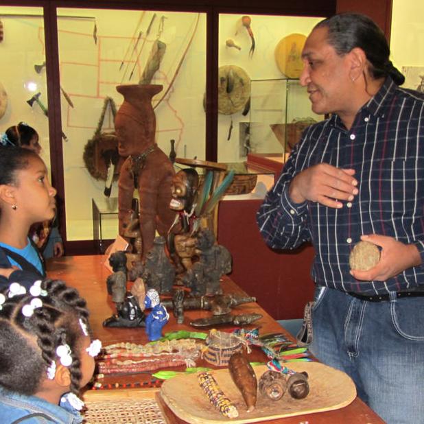 Three children listen with interest to a man speaking with them across a table of cultural items in front of anthropology gallery cases. 