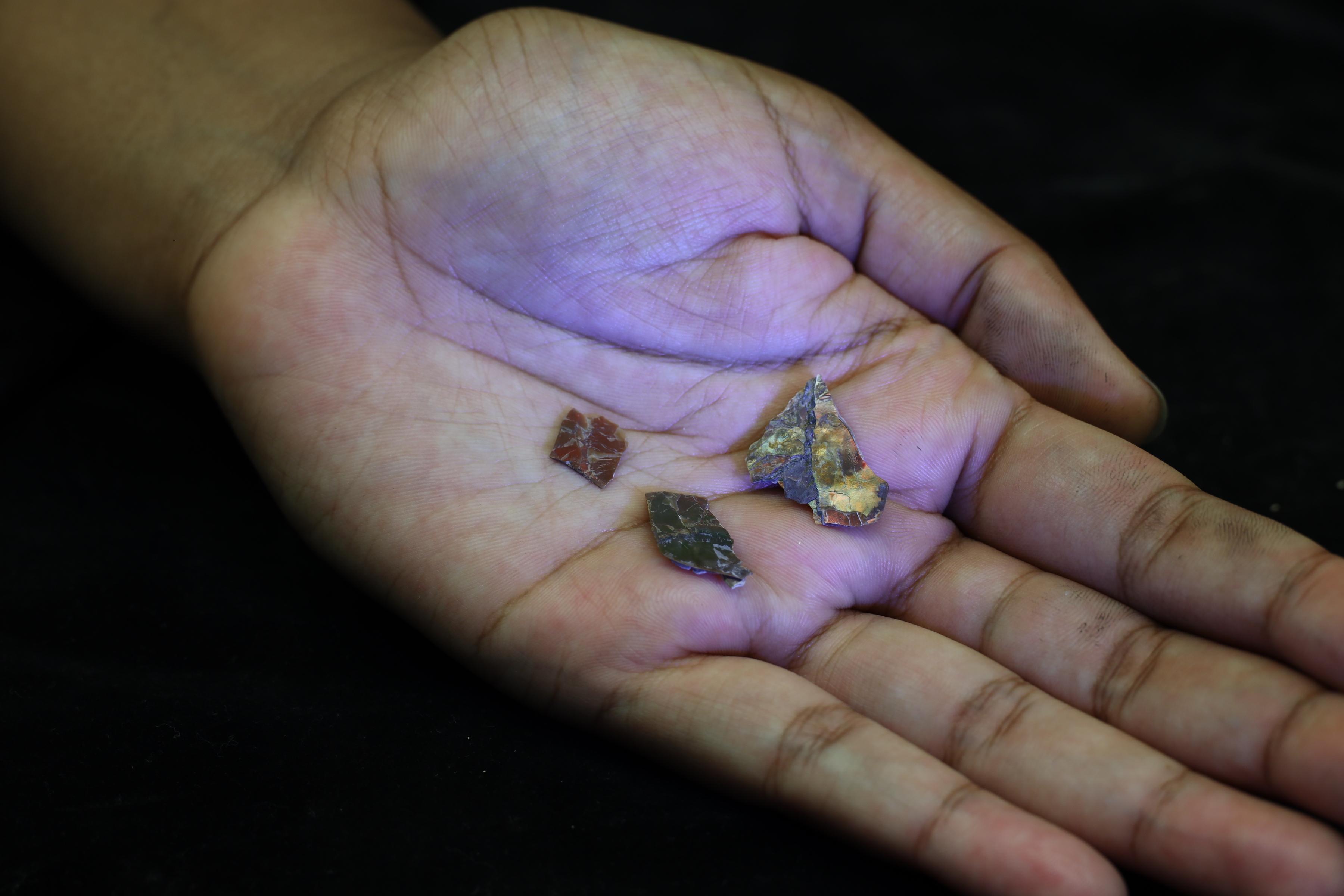 A dark-skinned hand holds three small fractured shards of stone with reflective and red-green surfaces